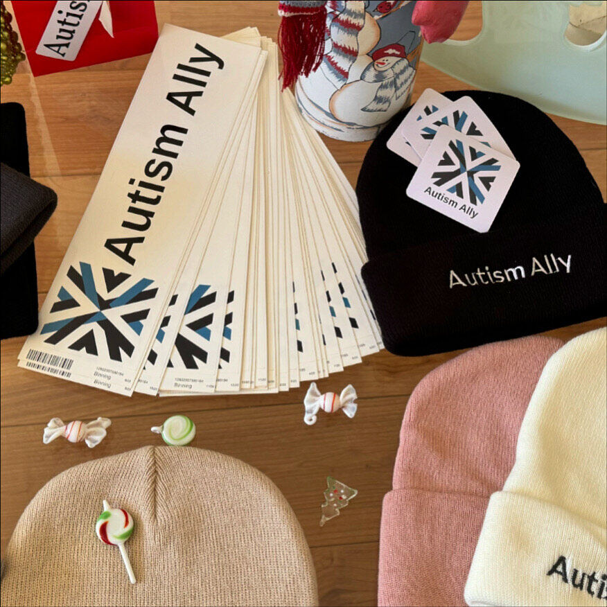 Autism Ally beanies in black, pink, cream and beige laid out with bumper stickers and square stickers.