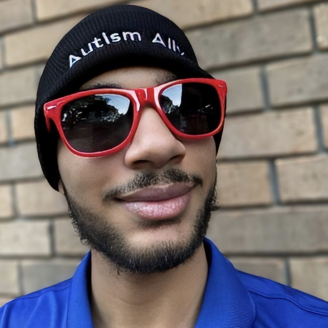 A man in a black Autism Ally beanie and red sunglasses against a brick wall.