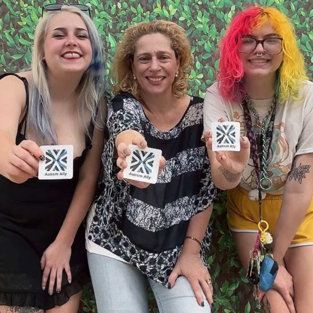 Three people holding Autism Ally stickers in front of a green hedge.